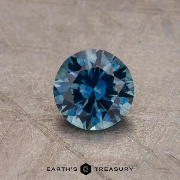 1.37-Carat Montana Sapphire (Heated) A close-up image of a round, 2.14-carat Robin's Egg Blue Montana Sapphire with intricate facets reflecting light, placed on a textured, gray surface. The text "EARTH'S TREASURY" and a small hexagonal logo are positioned at the bottom of the image.