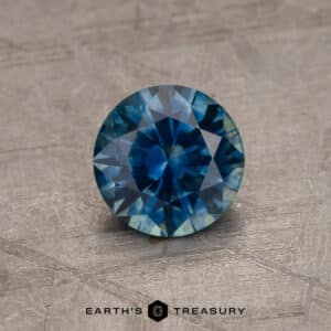 A round, faceted 2.14-Carat Robin's Egg Blue Montana Sapphire (Heated) is displayed against a textured gray background. The intricate cuts and reflections highlight its brilliance and clarity. The logo "Earth's Treasury" is positioned beneath the gemstone.