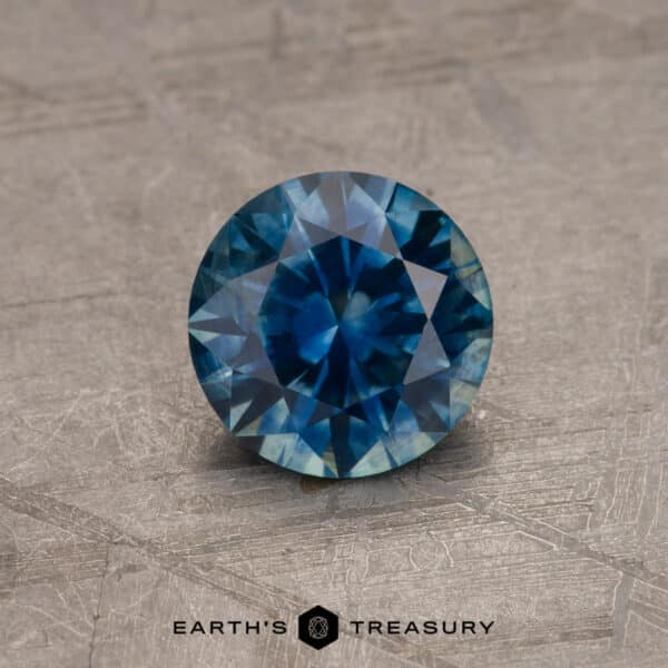 1.34-Carat Montana Sapphire (Heated) A round, faceted 2.14-Carat Robin's Egg Blue Montana Sapphire (Heated) is displayed against a textured gray background. The intricate cuts and reflections highlight its brilliance and clarity. The logo "Earth's Treasury" is positioned beneath the gemstone.