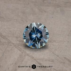 A brilliant-cut, round 0.81-carat aqua blue Montana sapphire with a striking blue color is displayed on a textured, beige surface. The text "Earth's Treasury" is visible at the bottom of the image.