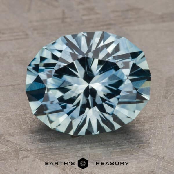 4.14-Carat Montana Sapphire (Heated) A 2.14-carat Robin's Egg Blue Montana Sapphire (heated) is centered on a textured gray surface. The gem exhibits a vivid sparkle and intricate cuts, showcasing various shades of blue. Below the gemstone, "Earth's Treasury" is printed in black text.