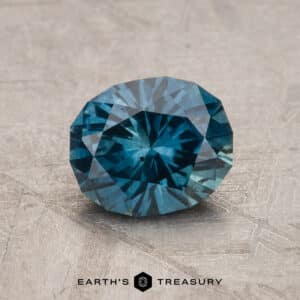 A 2.14-carat Robin's Egg Blue Montana Sapphire (heated) sits on a textured gray surface with intricate patterns. The gem's brilliant cut and vivid teal hue capture the light, creating a radiant display of color. The text "Earth's Treasury" is visible at the bottom of the image.