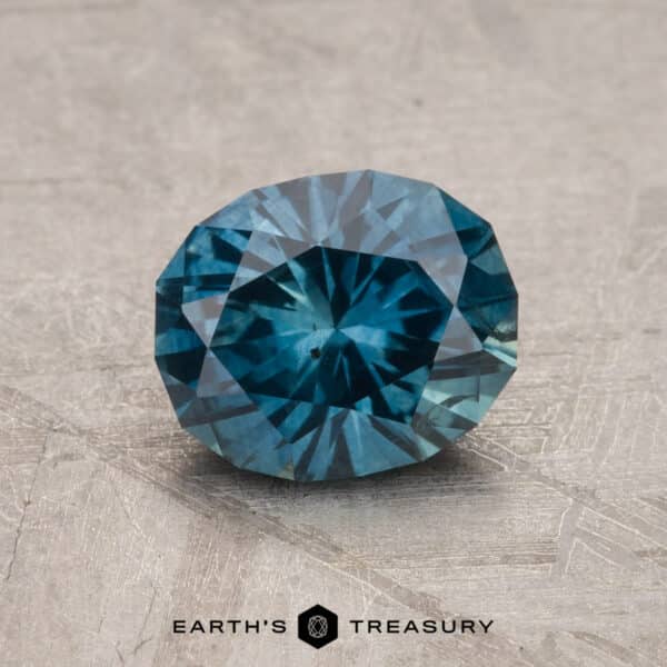 1.68-Carat Montana Sapphire (Heated) A 2.14-carat Robin's Egg Blue Montana Sapphire (heated) sits on a textured gray surface with intricate patterns. The gem's brilliant cut and vivid teal hue capture the light, creating a radiant display of color. The text "Earth's Treasury" is visible at the bottom of the image.