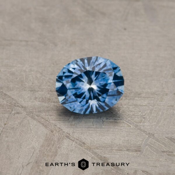 0.70-Carat Montana Sapphire (Heated) A brilliant blue gemstone, the 2.14-Carat Robin's Egg Blue Montana Sapphire (Heated), sits on a textured, beige surface. The gem is faceted and catches the light, showcasing its sparkling, vivid blue color. The words "EARTH'S TREASURY" are visible at the bottom of the image.