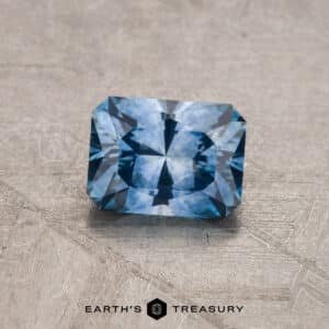 A 2.14-carat, rectangular-cut Robin's Egg Blue Montana Sapphire (heated) lies on a textured gray surface. The gemstone has intricate facets, reflecting light and creating a pattern of varying blue shades. Below the gemstone, the text reads "EARTH'S TREASURY" accompanied by a small geometric logo.