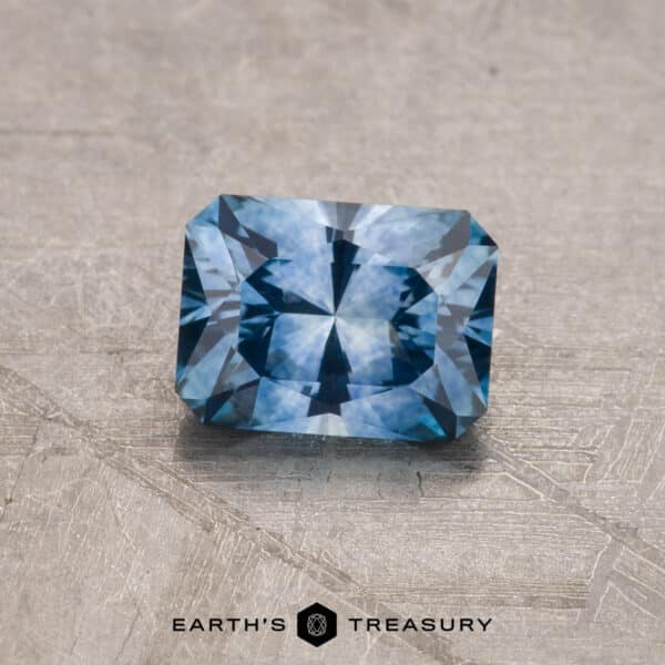 0.95-Carat Montana Sapphire (Heated) A 2.14-carat, rectangular-cut Robin's Egg Blue Montana Sapphire (heated) lies on a textured gray surface. The gemstone has intricate facets, reflecting light and creating a pattern of varying blue shades. Below the gemstone, the text reads "EARTH'S TREASURY" accompanied by a small geometric logo.