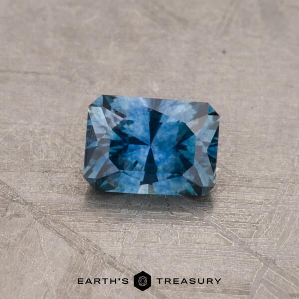 A captivating 2.14-carat Robin's Egg Blue Montana Sapphire, shaped in a rectangular cut with faceted edges, is set against a textured gray surface. The sapphire displays beautiful variations in blue hues and internal reflections. Beneath the gemstone is the text "EARTH'S TREASURY" accompanied by a small geometric logo.
