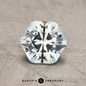A sparkling 2.14-carat robin's egg blue Montana sapphire with a hexagonal shape and numerous facets is placed on a textured surface. The gem has a pale light-blue color and is positioned centrally above the text "Earth's Treasury," accompanied by a small, black hexagonal logo.