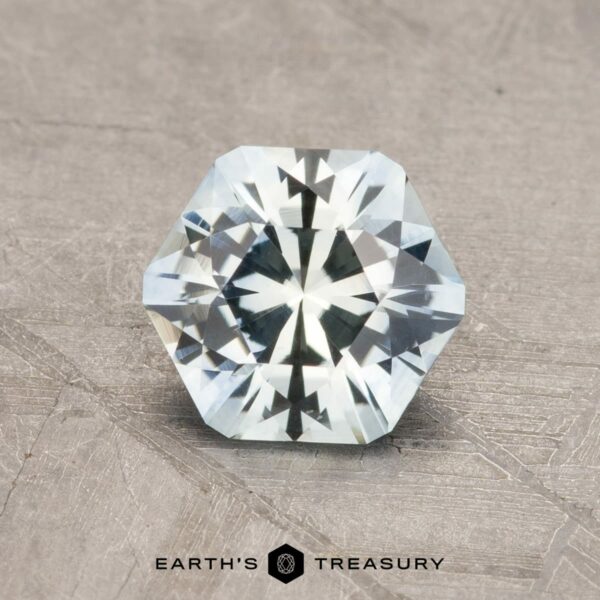1.17-Carat Montana Sapphire (Heated) A sparkling 2.14-carat robin's egg blue Montana sapphire with a hexagonal shape and numerous facets is placed on a textured surface. The gem has a pale light-blue color and is positioned centrally above the text "Earth's Treasury," accompanied by a small, black hexagonal logo.
