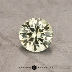 A round, faceted gemstone with a light yellow tint is displayed against a textured gray background. The gemstone's intricate cuts highlight its brilliance and sparkle. The text "2.14-Carat Robin's Egg Blue Montana Sapphire (Heated)" is visible at the bottom of the image under "EARTH'S TREASURY.