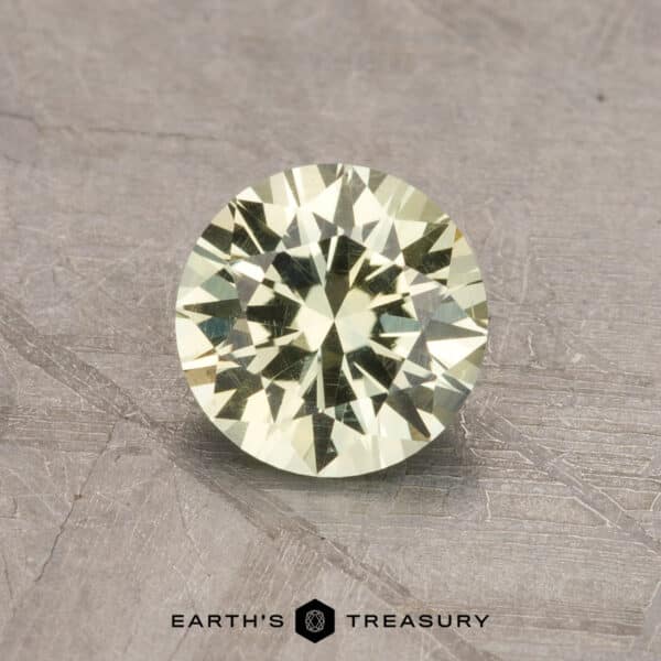 1.06-Carat Montana Sapphire (Heated) A round, faceted gemstone with a light yellow tint is displayed against a textured gray background. The gemstone's intricate cuts highlight its brilliance and sparkle. The text "2.14-Carat Robin's Egg Blue Montana Sapphire (Heated)" is visible at the bottom of the image under "EARTH'S TREASURY.