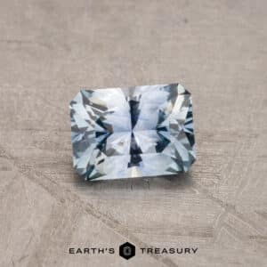 A radiant 2.14-carat Robin's Egg Blue Montana Sapphire, heated for enhancement, displayed on a textured surface, catching light and showcasing its blue hues and intricate facets. The text "EARTH'S TREASURY" is visible below the gemstone.