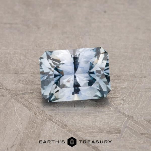 0.80-Carat Montana Sapphire (Heated) A radiant 2.14-carat Robin's Egg Blue Montana Sapphire, heated for enhancement, displayed on a textured surface, catching light and showcasing its blue hues and intricate facets. The text "EARTH'S TREASURY" is visible below the gemstone.