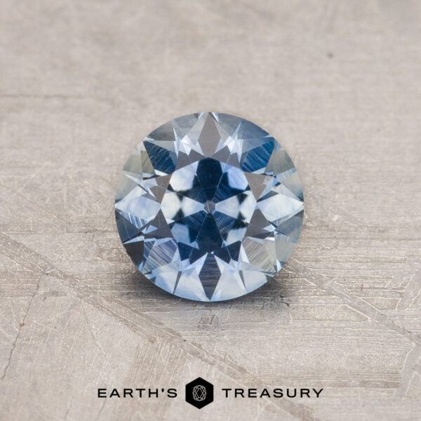 0.82-Carat Montana Sapphire (Heated) A 2.14-carat Robin's Egg Blue Montana Sapphire (heated) sits on a textured gray surface. The gemstone's intricate facets reflect light, showcasing its clarity and brilliance. Below the gem, the text reads "EARTH'S TREASURY" with a small emblem in between the words.