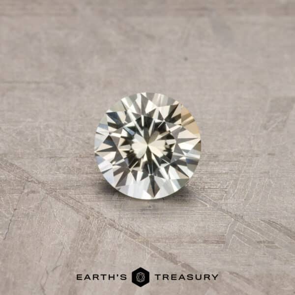 0.78-Carat Montana Sapphire (Heated) A brilliant-cut round gemstone, the 2.14-Carat Robin’s Egg Blue Montana Sapphire (Heated), displayed on a wooden surface. The gem exhibits a high level of sparkle and clarity. The text "EARTH'S TREASURY" is visible at the bottom of the image.