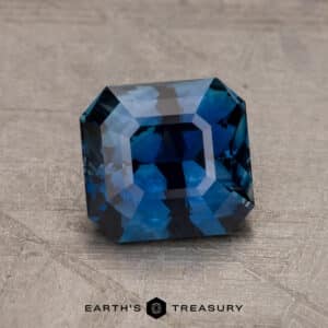 A 2.14-carat Robin's Egg Blue Montana Sapphire with square-cut and beveled edges is showcased on a textured gray surface. The sapphire reflects light, highlighting its facets and cuts. Below the gemstone, "Earth's Treasury" is written in black text.