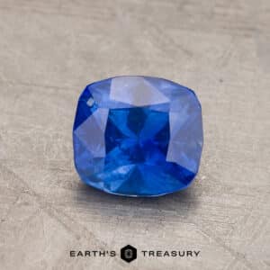 A radiant 2.14-carat Robin's Egg Blue Montana Sapphire, cushion-cut and heated, is showcased against a gray textured background. The bottom center of the image includes the text "EARTH'S TREASURY" with a hexagonal logo between the words.