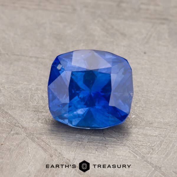 1.41-Carat Sapphire (Heated) A radiant 2.14-carat Robin's Egg Blue Montana Sapphire, cushion-cut and heated, is showcased against a gray textured background. The bottom center of the image includes the text "EARTH'S TREASURY" with a hexagonal logo between the words.