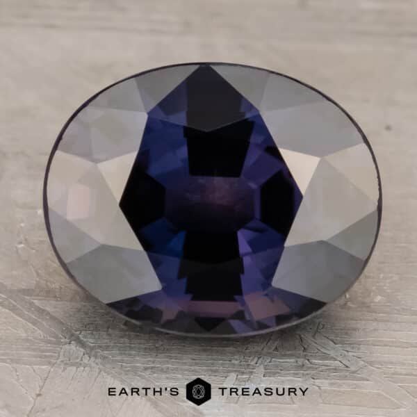 Incandescent A 3.10-carat bluish violet to purple color-change sapphire, faceted in an oval shape, is showcased against a textured, grey background. The gem's intricate cutting creates a brilliant display of reflective surfaces. Text at the bottom reads "Earth's Treasury.