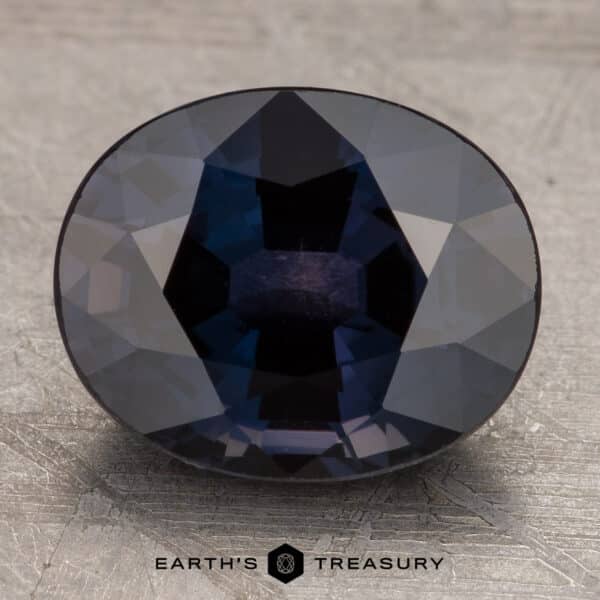 Daylight A 3.10-carat, faceted, oval-shaped Bluish Violet to Purple Color-Change Sapphire is displayed against a textured, silver-gray background. The gemstone displays various shades of blue under the lighting, and the text "EARTH'S TREASURY" is printed at the bottom center.