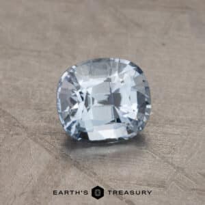 A 2.14-carat Robin's Egg Blue Montana Sapphire with a cushion cut shape is displayed on a textured surface. The clarity and facets of the gem are highlighted by the lighting. At the bottom of the image, the text reads "EARTH'S TREASURY" with a small geometric logo.
