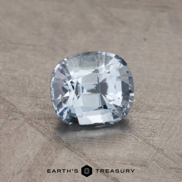 1.24-Carat Sapphire A 2.14-carat Robin's Egg Blue Montana Sapphire with a cushion cut shape is displayed on a textured surface. The clarity and facets of the gem are highlighted by the lighting. At the bottom of the image, the text reads "EARTH'S TREASURY" with a small geometric logo.