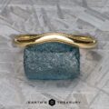 A stunning "Narcissus" Band in 18k yellow gold, featuring a textured edge, is elegantly displayed on a serene blue stone atop a patterned grey surface. Below it, the text reads "Earth's Treasury," evoking the elegance and harmony akin to Narcissus Band's finest tunes.