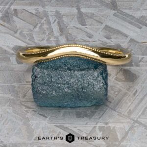 A stunning "Narcissus" Band in 18k yellow gold, featuring a textured edge, is elegantly displayed on a serene blue stone atop a patterned grey surface. Below it, the text reads "Earth's Treasury," evoking the elegance and harmony akin to Narcissus Band's finest tunes.