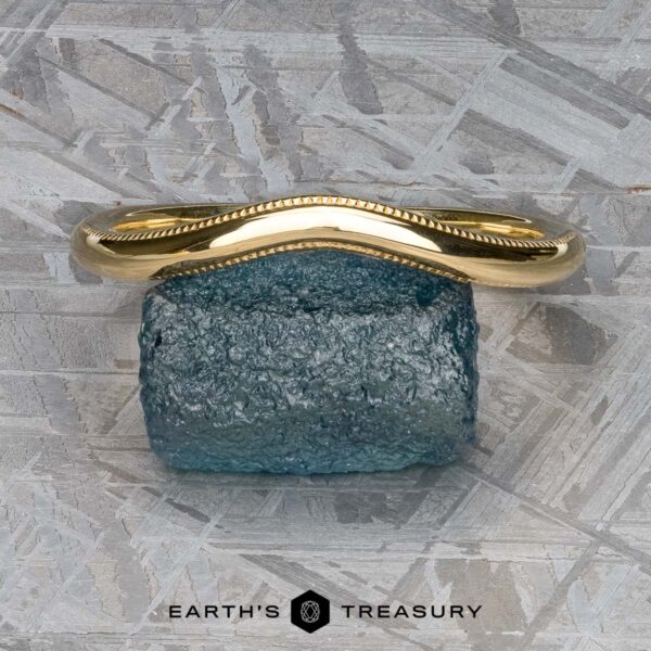 The "Narcissus" Band in 18k yellow gold A stunning "Narcissus" Band in 18k yellow gold, featuring a textured edge, is elegantly displayed on a serene blue stone atop a patterned grey surface. Below it, the text reads "Earth's Treasury," evoking the elegance and harmony akin to Narcissus Band's finest tunes.