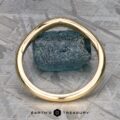 An 18k yellow gold "Narcissus" band elegantly encircles a raw blue gemstone, resting gracefully on a textured gray surface. Text at the bottom reads "Earth's Treasure," evoking the beauty found in nature's own symphony.