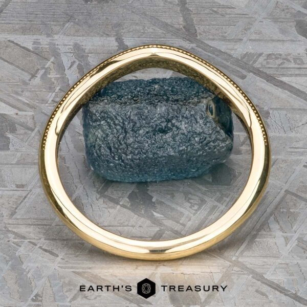 The "Narcissus" Band in 18k yellow gold An 18k yellow gold "Narcissus" band elegantly encircles a raw blue gemstone, resting gracefully on a textured gray surface. Text at the bottom reads "Earth's Treasure," evoking the beauty found in nature's own symphony.