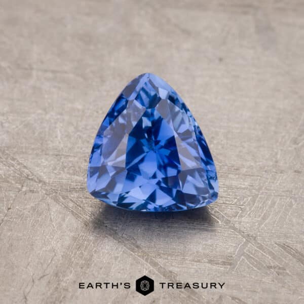 1.41-Carat Ceylon Sapphire A vibrant royal blue, trillion-cut 1.53-carat Ceylon sapphire rests on a textured beige surface. The gem's facets catch the light, showcasing its clarity and depth of color. The photo features "Earth's Treasury" text at the bottom.