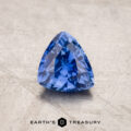 A vibrant royal blue, trillion-cut 1.53-carat Ceylon sapphire rests on a textured beige surface. The gem's facets catch the light, showcasing its clarity and depth of color. The photo features "Earth's Treasury" text at the bottom.