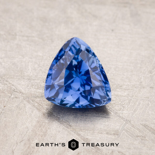 A vibrant royal blue, trillion-cut 1.53-carat Ceylon sapphire rests on a textured beige surface. The gem's facets catch the light, showcasing its clarity and depth of color. The photo features "Earth's Treasury" text at the bottom.