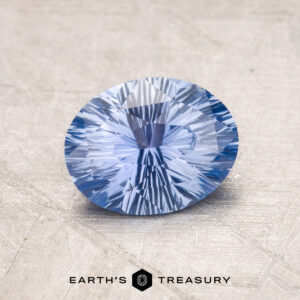 A faceted oval-cut blue 1.38-Carat Ceylon Sapphire displayed on a textured surface with the text "EARTH'S TREASURY" at the bottom.