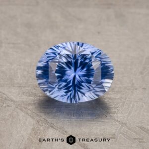 A 1.53-carat royal blue Ceylon sapphire with an oval cut and intricate, symmetrical facets is placed on a textured, light grey surface. Below the gemstone, the text "Earth's Treasury" is visible.