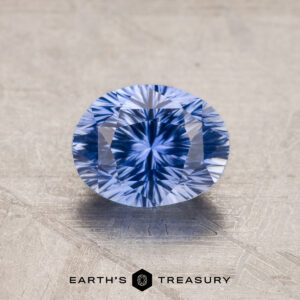A 1.53-carat royal blue Ceylon sapphire with an oval cut and intricate, symmetrical facets is placed on a textured, light grey surface. Below the gemstone, the text "Earth's Treasury" is visible.
