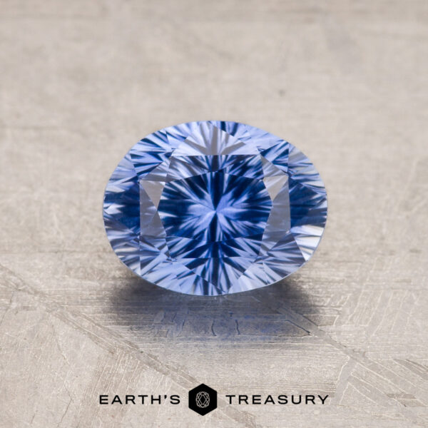 1.73-Carat Ceylon Sapphire A 1.53-carat royal blue Ceylon sapphire with an oval cut and intricate, symmetrical facets is placed on a textured, light grey surface. Below the gemstone, the text "Earth's Treasury" is visible.