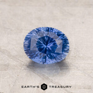 A 1.53-carat Royal Blue Ceylon Sapphire (heated) with intricate cuts is displayed on a textured, beige surface. The gemstone's vivid blue color and precise facets reflect light brilliantly. The Earth's Treasury logo is present at the bottom of the image.
