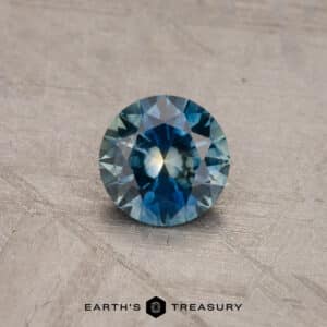A 1.53-carat Royal Blue Ceylon Sapphire (heated) with a mix of deep blue and light greenish-blue hues, displayed on a textured gray surface. The gemstone exhibits high clarity and intricate facets. The text "Earth's Treasury" appears at the bottom of the image.