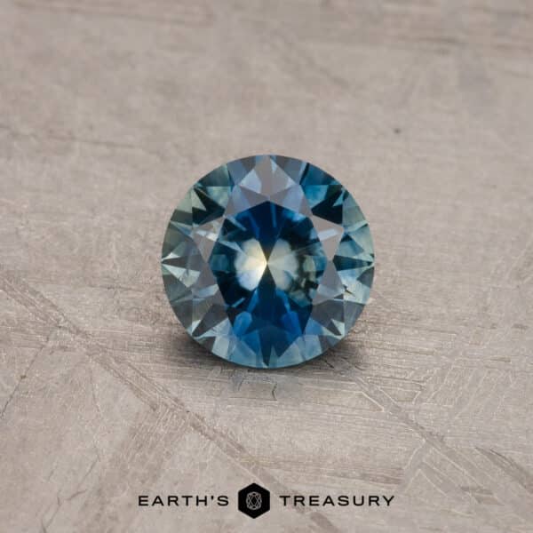 0.95-Carat Montana Sapphire (Heated) A 1.53-carat Royal Blue Ceylon Sapphire (heated) with a mix of deep blue and light greenish-blue hues, displayed on a textured gray surface. The gemstone exhibits high clarity and intricate facets. The text "Earth's Treasury" appears at the bottom of the image.