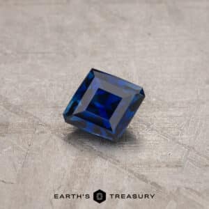 A 1.53-carat Royal Blue Ceylon Sapphire, precisely cut into a square shape, rests on a textured grey surface. The background is adorned with subtle geometric patterns, and the text "EARTH'S TREASURY" is prominently displayed at the bottom of the image.