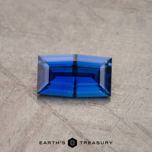 0.91-carat Ceylon sapphire (Heated) A brightly lit, meticulously faceted 1.53-carat Royal Blue Ceylon Sapphire (heated) sits on a textured, light-colored surface. The gem's geometric shape features clean, sharp lines and angles, showcasing its brilliant cut and multiple facets. Below the gem is the text "Earth's Treasury.