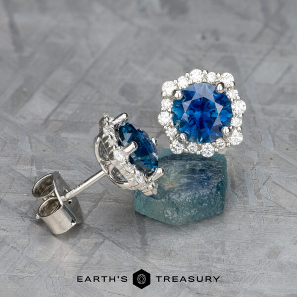 EA-MS-521 Pair of silver halo earrings (EA-MS-521) featuring round Montana sapphire centers surrounded by clear stones, displayed on a rough blue gemstone with the "Earth's Treasury" logo below.