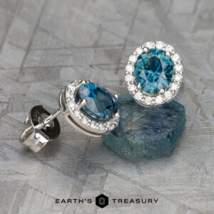 EA-MS-508: Silver Halo Earrings featuring round Montana Sapphire gemstones encircled by small clear stones, shown on a rough blue stone with a geometric background.