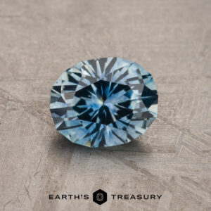 A brilliantly cut 1.30-carat Sky Blue Montana Sapphire (Heated) rests on a textured surface. "Earth's Treasury" logo and text appear at the bottom.