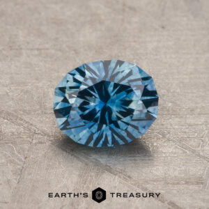 A 1.18-Carat Teal Blue Montana Sapphire (Heated) with an oval-faceted cut is showcased on a textured gray background with the Earth's Treasury logo at the bottom.