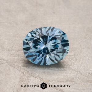 A 1.10-carat aqua blue Montana Sapphire (heated), faceted in an oval shape, rests on a textured gray surface with "EARTH'S TREASURY" appearing at the bottom.