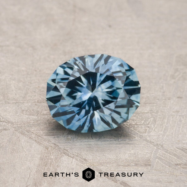 1.10-Carat Aqua Blue Montana Sapphire (Heated) A 1.10-carat aqua blue Montana Sapphire (heated), faceted in an oval shape, rests on a textured gray surface with "EARTH'S TREASURY" appearing at the bottom.