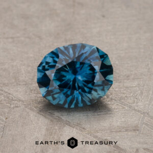 The 1.10-Carat Peacock Blue Montana Sapphire (Heated) is displayed on a textured, light-toned surface with "EARTH'S TREASURY" written at the bottom.